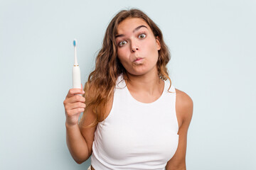 Young caucasian woman holding electric toothbrush isolated blue background shrugs shoulders and open eyes confused.
