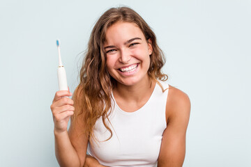 Young caucasian woman holding electric toothbrush isolated blue background laughing and having fun.