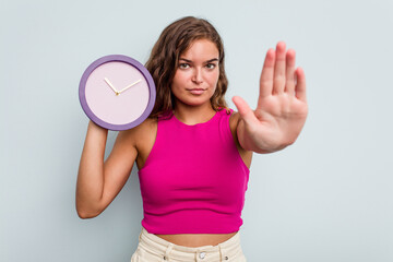 Young caucasian woman holding a clock isolated on blue background standing with outstretched hand showing stop sign, preventing you.