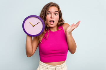 Young caucasian woman holding a clock isolated on blue background surprised and shocked.