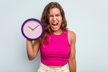 Young caucasian woman holding a clock isolated on blue background screaming very angry and aggressive.