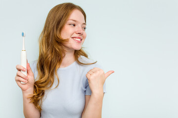 Young caucasian woman holding electric toothbrush isolated on blue background points with thumb finger away, laughing and carefree.