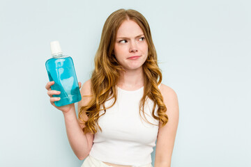 Young caucasian woman holding a mouthwash isolated on blue background confused, feels doubtful and...