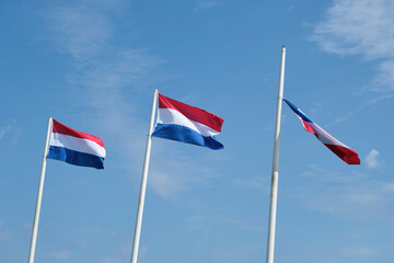 Three red white blue dutch flags on flagpoles in strong wind against a blue sky with large clouds. One of them is loose.	
