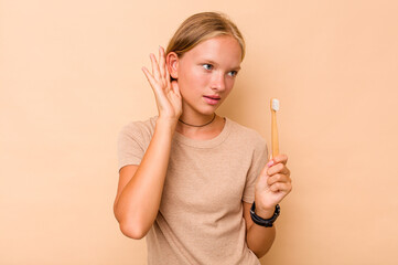 Caucasian teen girl brushing teeth isolated on beige background trying to listening a gossip.