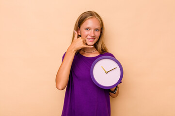 Little caucasian girl holding a clock isolated on beige background showing a mobile phone call gesture with fingers.