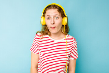 Young caucasian woman wearing headphones isolated on blue background shrugs shoulders and open eyes confused.