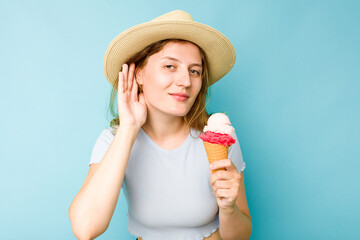 Young caucasian woman holding an ice cream isolated a blue background trying to listening a gossip.