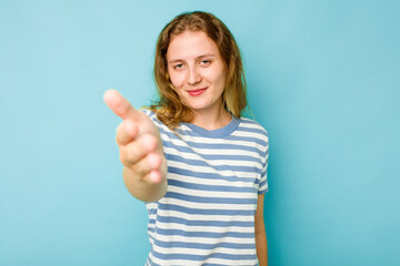 Young caucasian woman isolated on blue background stretching hand at camera in greeting gesture.