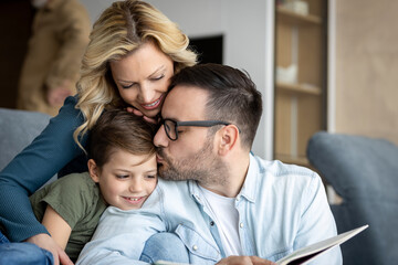 Obraz premium A loving parents sits on the couch at home and reads a storybook to there's elementary school age son. The child is snuggle between mother and father and is smiling while looking at the book. 