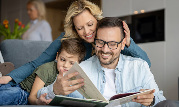 A loving parents sits on the couch at home and reads a storybook to there's elementary school age son. The child is snuggle between mother and father and is smiling while looking at the book.