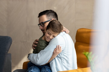 A loving parent sits on the sofa at home and hugging his son. The child is sitting in fathers lap and enjoying moment with his father