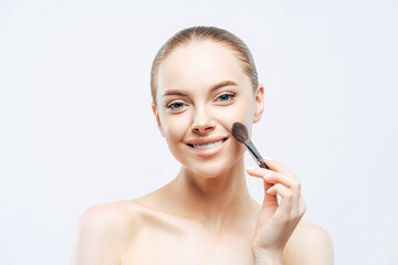 Fototapeta premium Adorable young brunette woman applies cosmetic tonal foundation with beauty brush, smiles tenderly, has healthy skin, well cared body, isolated on white background. Women, skin care, makeup concept