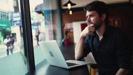 Concerned pensive young entrepreneur man in front of laptop computer seated by window at cafe co working remote workplace