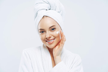 Charming relaxed beauty woman applies face cream, cares about complexion, touches cheek with hand, smiles gently at camera, dressed in bath robe, wrapped towel on washed hair, isolated on white