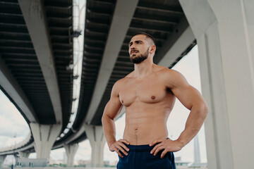 Athletic male fitness model keeps hands on waist poses with muscular perfect naked body, looks seriously into distance, does sport exercises outdoor, focused somewhere, goes in for sport regularly