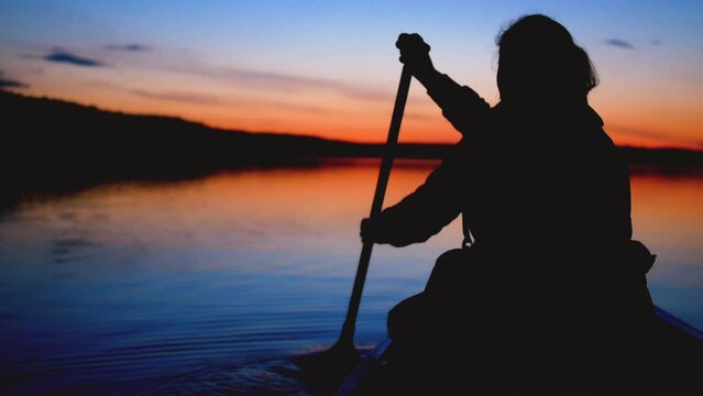 Static view of a woman paddling a canoe on lake. Nighttime atmosphere with setting sunlight. Sweden