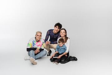 happy lesbian woman sitting with toddler boy near gay man and queer person on grey background.