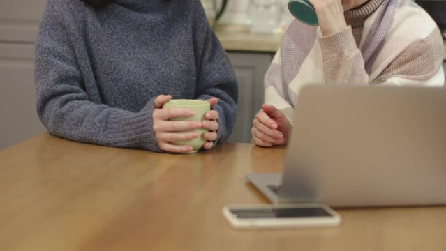 two woman warm clothes sit kitchen hold cup hot beverage talking drinking tea laptop computer smartphone on table. concept female family friends gossiping together at home indoors. cold apartment 