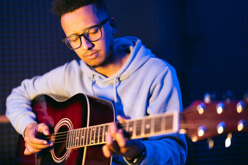 Obraz premium Young afro musician wearing glasses playing acoustic guitar, on dark background