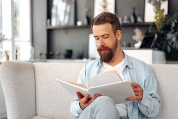 Young bearded handsome man reading book on couch at home