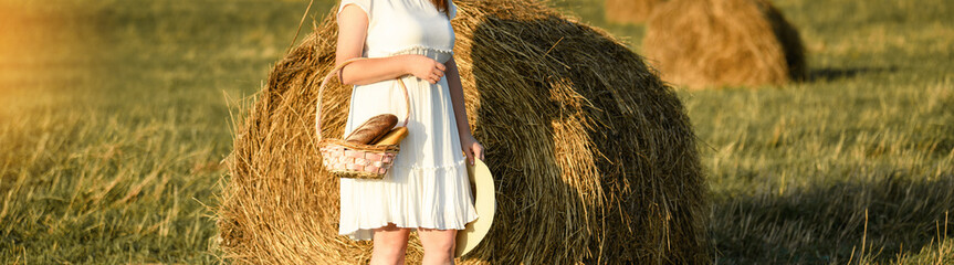 Girl and bread. A woman in a white dress stands with a basket of bread in the background of a field with groans of hay. Harvest concept. Banner