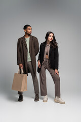full length of stylish african american in autumnal coat posing with paper bags near woman on grey.