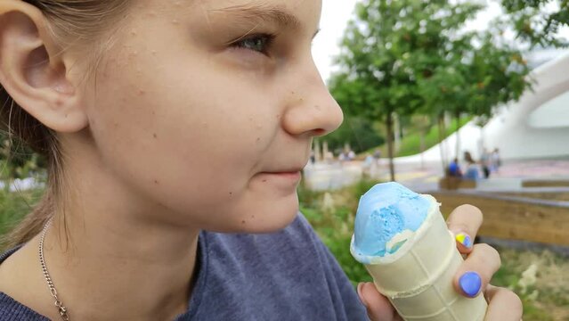 Beautiful teen girl eating ice cream. Carefree childhood and children's joys. Turquoise ice cream in a waffle cone or cup. A delicious treat on a hot summer day in a city public park. Close-up