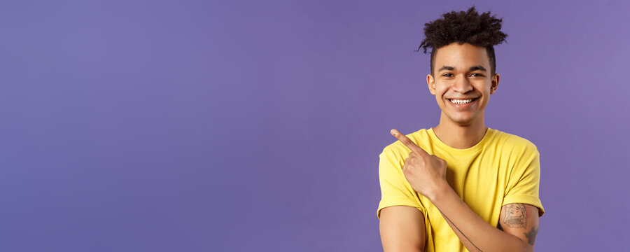 Close-up portrait of enthusiastic, happy young hipster male with dreads, beaming smile and pointing finger upper left corner, present cool product, introduce something really good, purple background