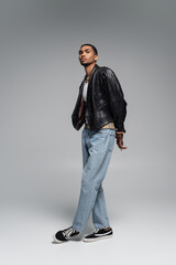 full length of young african american man in jeans and leather black jacket posing on grey.