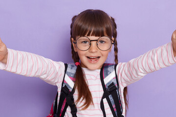 Beautiful smiling Caucasian little girl with glasses and with school bag is back to school, posing...