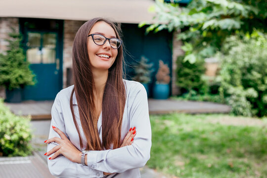 A young student girl in glasses smiles with folded arms dressed in a shirt against the background of the facade of the building