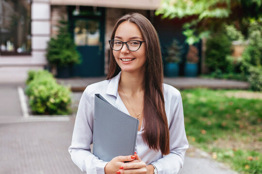 a girl student in glasses holds a folder and smiles against the background of the facade of the building