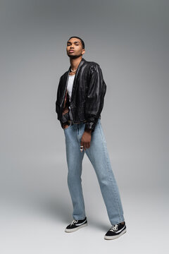full length of young african american man in jeans and leather black jacket on grey.