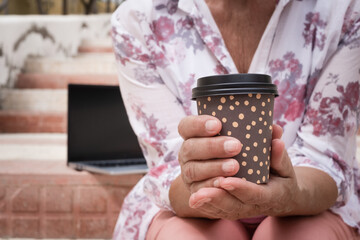 Close-up on senior woman's hands holding a takeaway coffee cup while sitting outdoors on a stairway in the city close to her laptop