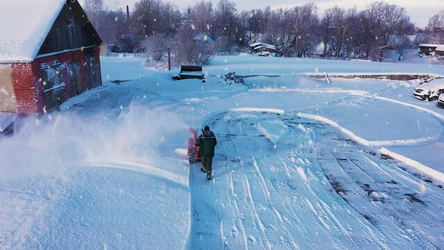 Man plowing snow with hand machine near old rural farm building, aerial view
