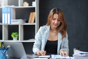 Beautiful Asian woman sitting in the office using a laptop. Happy business woman smiling and enjoying her work and taking note on a notepad.