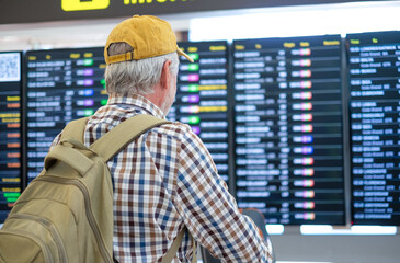 Old senior man in airport pushing luggage cart looking at timetable schedule to check his flight departure. Traveler concept people with backpack and suitcases.