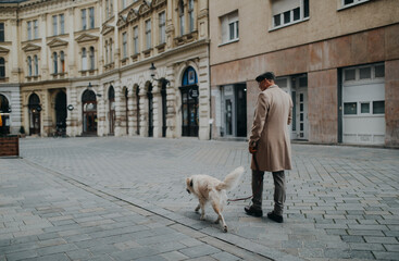 Side view of elegant senior man with take away coffee walking his dog outdoors in city in winter.