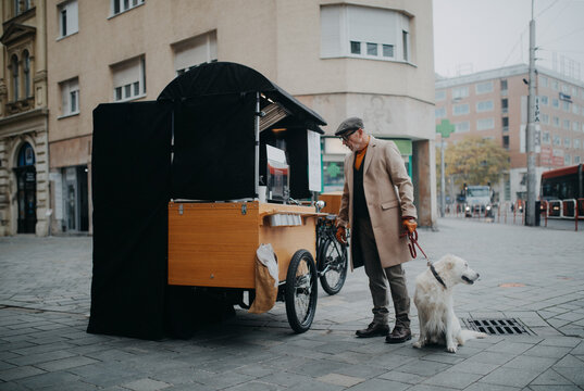 Elegant senior man walking his dog and buiyng street food outdoors in city.