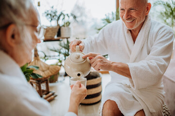 Senior couple in bathrobes enjoying time together in their living room, drinking hot tea, calm and hygge atmosphere.