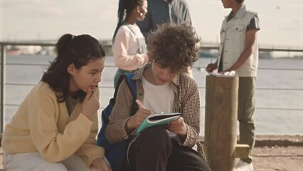 Medium slowmo of multiethnic classmates chatting while doing school project on alternative energies outdoors at sunny quay with their teacher standing with windmill model in background