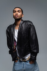 Fototapeta premium young african american man in jeans and black jacket posing with hands in pockets isolated on grey.