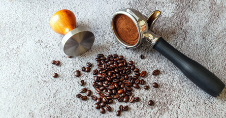 Equipment in a coffee shop of barista coffee tool portafilter with tamper and dark roasted coffee beans on gray background