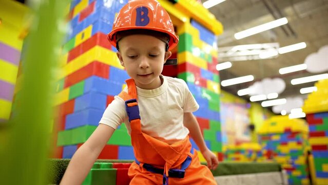 Handsome little boy dressed in a construction helmet plays big colored blocks. The child plays builder on the playground. Educational games for children.
