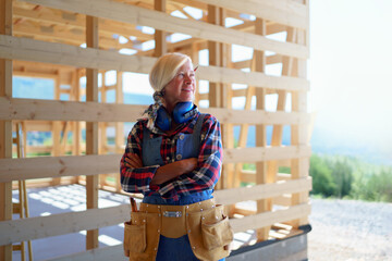 Senior woman in working clothes posing outdoor of her unfinished ecological sustainable wooden eco house. Concept of active independent women and seniors, eco house and healthy lifestyle.