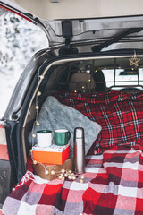 Trunk of car is decorated for celebration of Christmas and New Year in snowy winter forest. Mug and thermos for drinking hot tea. Cozy red plaid and gift box. White picnic in woods.