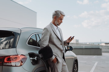 Businessman holding smartphone while charging car at electric vehicle charging station, closeup.