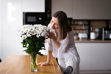 Happy and joyful young woman in white arranging white flowers at home in the kitchen