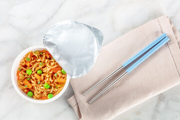 Ramen cup, instant soba noodles in a plastic cup with vegetables, with chopsticks, overhead flat lay shot on a white marble table